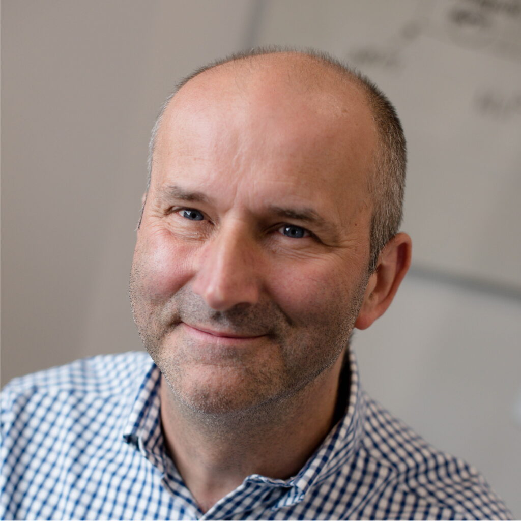 A photo of Andy's head and shoulders. He is a white male with shortly cropped greying hair and blue eyes. Andy is smiling and looking into the camera.